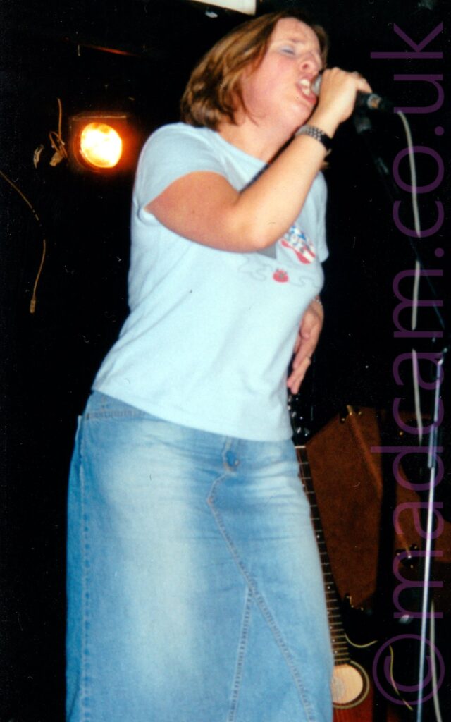A woman with chin-length blonde hair, wearing a blue t-shirt and a long blue denim skirt, singing on a stage. She is holding a microphone on a stand to her face with her right hand, her left hand held down by the side of her waist. Black walls and a yellow stage light fill the background.