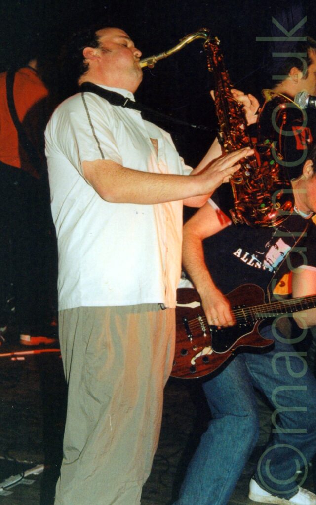 A man with short dark hair, standing facing to the right, wearing a white shirt and beige trousers, playing a gold-coloured saxophone attached to a black strap around his neck. In the background on the right, a man with short, brown-ish hair and wearing a dark blue t-shirt and a pair of blue jeans is slightly crouched over, playing a red electric guitar, with another man behind him with only the side of his face visible. A man with his back to the camera, wearing a red top, is in the background on the left. Black walls fill the rest of the frame.