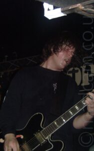 A man, with chin-length dark hair and wearing a black sweatshirt, standing facing to the right, playing a black electric guitar. His face and hair are dripping with sweat. Black walls and ceiling fill the background, with a metal frame stretching across the top of the back wall, holding up a black banner with the white letters "OP" visible.
