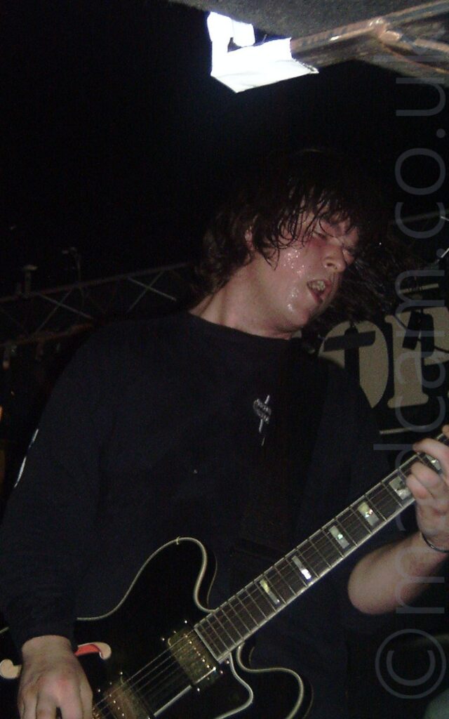 A man, with chin-length dark hair and wearing a black sweatshirt, standing facing to the right, playing a black electric guitar. His face and hair are dripping with sweat. Black walls and ceiling fill the background, with a metal frame stretching across the top of the back wall, holding up a black banner with the white letters "OP" visible.