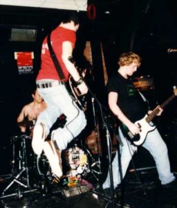 3 men performing on a stage as part of a band. In the middle of the frame is a man with short dark hair, wearing a red t-shirt and faded blue jeans, leaping high into the air while playing a black electric guitar. On the right is a man with light-coloured, curly hair, wearing a black t-shirt with faded blue jeans, and playing a black bass guitar with a white plate on the front, leaning back as he plays At the back is a shirt-less man sitting behind a black and chrome drum kit. Black walls fill the rest of the frame.