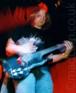 Low shot of a man with floppy blond hair, wearing a black t-shirt with an indistinct white logo on the chest and a blue pair of jeans, playing a black electric guitar while staring moodily at the camera. A long exposure shows movement in the guitar and his hands, with a streak of red light in the background behind him. Red and black walls, and a black ceiling, fill the rest of the frame.