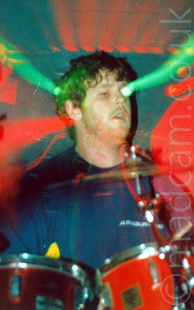 A man with curly, short brown hair, wearing a dark blue t-shirt, sitting behind a red and chrome drum kit. A flash has frozen him in place, while a long exposure and a snap zoom out has created streaks of green and red light centred on his head, with additional blooms of red light to either side of him, and on the black wall behind him.