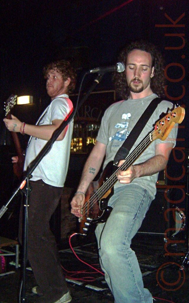 Two men performing on a stage as part of a rock band. On the left is a man with curly brown hair, with stubble on his face, showing a rather surprised expression. He is wearing a white t-shirt and brown trousers, and playing a brown electric guitar. On the right is a man with shoulder-length, dark, curly hair, with a thin, dark goatee beard and moustache. He is wearing a grey t-shirt with a white and blue logo featuring some of the cars from the Wacky Races cartoon show, and a faded blue pair of jeans, and is playing a black bass guitar with an orange sunburst on the front. Speaker cabinets are on the floor against the back wall, with a mirror hanging on the wall in the background, with the letters "URCH" visible in a curve towards the top. Black walls and ceiling fill the rest of the background.