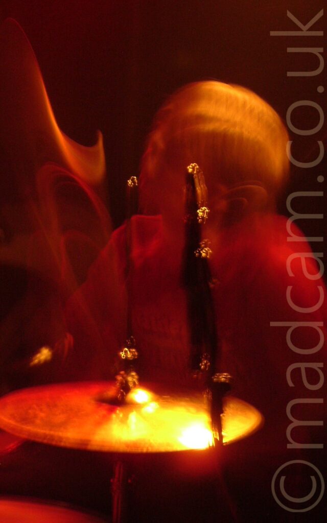A long exposure shot of a man with long hair playing a drum kit, his arms and head mostly smears of red and orange light .A cymbal bathed in yellow, orange, and red light is at the bottom of the frame.
