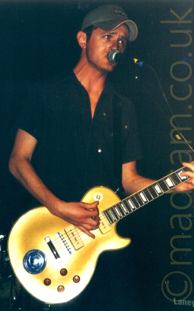 A man wearing a grey baseball cap and a black shirt, playing a metallic yellow electric guitar and singing into a microphone on a stand in front of him. Black walls and ceiling fill the background.