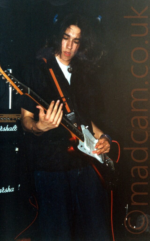 A man with long, dark hair flowing over his shoulders, wearing a black shirt over a white t-shirt, and dark blue jeans. He is playing a black electric guitar with a white face plat, and doing so left-handed, with his guitar strap over his right shoulder. A stack of Marshall amps is in the background on the left, with black walls and ceiling filling the rest.