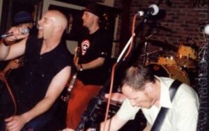 Three men performing on a stage as part of a band. On the left is a man with a shaven head, wearing a sleeveless black shirt and blue jeans, singing into a microphone held in his right hand, it's red cord wrapped around his right wrist. In the middle, at the back, is a man wearing a black pork pie hat, a black t-shirt with a white star in the middle of red and white circles, and bright red trousers, playing a bass guitar. On the right, slightly hunched over, is a man with short dark hair, wearing a short white shirt with a black guitar strap over his right shoulder. A microphone of a stand is over his head. A yellow and chrome drum kit can be seen at the back of the frame, in front of a brown brick wall.