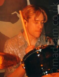 A man with floppy, ear-length blond hair, and wearing a sparkly silver shirt, sitting behind a black and chrome drum kit with a drum stick held in the air in his right hand. A large black, red, and white vinyl backdrop fills the background.