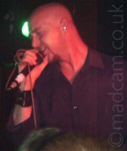 A man with a shaven head, wearing a black shirt, holding a microphone in his right hand to his mouth as he sings. The mic cord is wrapped around his fingers, and there is a black leather strap around his right wrist. Stage lights give his skin a red cast. The head of one of the people in the crowd is at the bottom of the frame, with a green light behind the singers head against the black walls.