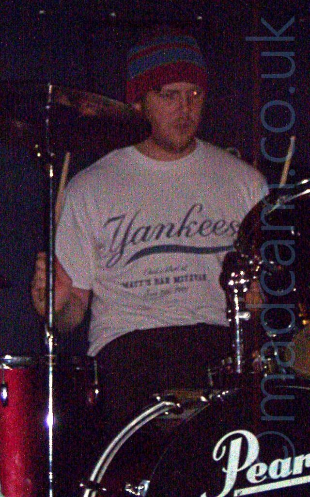 Rather grainy shot of a man with a shaggy little beard, wearing a red woollen hat with wide blue bands, a white t-shirt with a blue "Yankees" logo, and dark trousers, sitting behind a red drum kit, his arms raised slightly into the air, holding his drum sticks. Black walls fill the background.