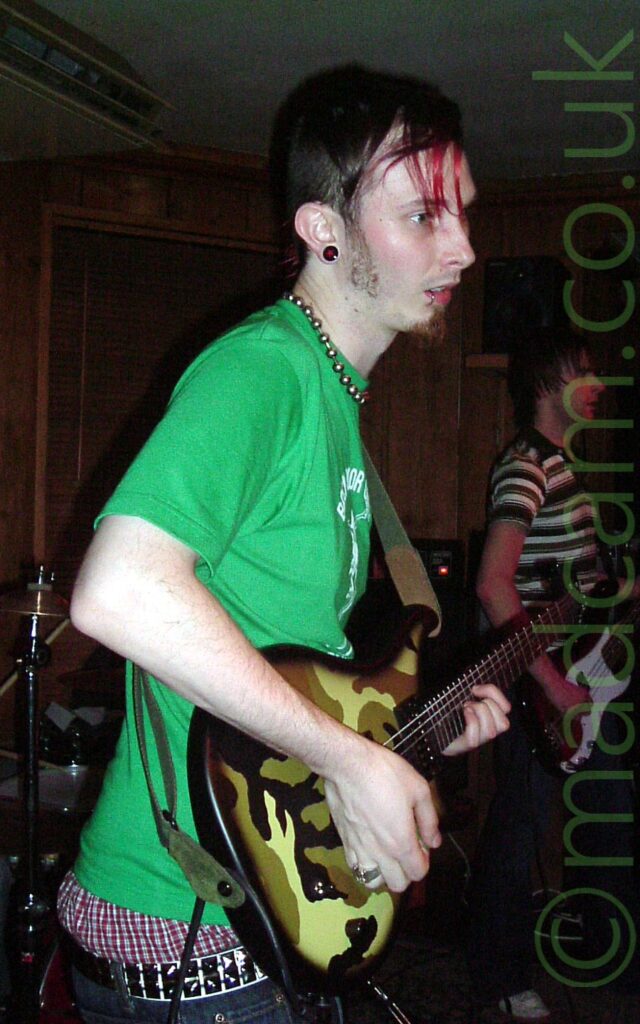 A man with mostly short dark hair, with long strands at the front, some dyed red, wearing a green t-shirt and low-hung blue jeans revealing red and black plaid boxer shorts, with a necklace made up of metal balls hanging around his neck. He is playing a black electric guitar with a green and brown camouflage scheme on the front. Another man, with lank, dank hair, and wearing a brown and white banded polo shirt, stands in the background on the right of the frame. Brown wooden walls and a white ceiling fill the rest of the frame.