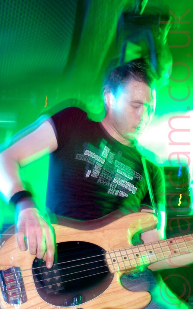 A man with short dark hair, wearing a black t-shirt with a white textual logo on the front, playing a light varnished wood bass guitar, leaning to his left. A flash from the camera has frozen him in place, while a long exposure has created smears of light where his body and instrument have moved, all bathed in green and blue light from the stage lights.