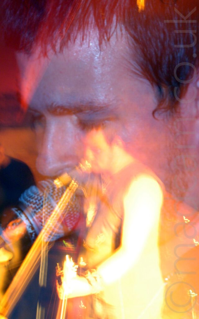 Close-up of the face of a man with short dark hair, singing into a microphone on a stand. There is a purple cast to his skin from the lights. Thanks to a long exposure and a snap zoom out, there is also a more distant shot of him, wearing a dark tank top and playing an electric guitar, while still singing into the mic, in various shades of yellow and orange.