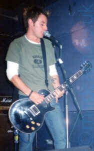 A man with short, slightly spiky hair, wearing a green t-shirt over a white long-sleeved t-shirt and a blue pair of jeans, playing a black electric guitar and singing in to a microphone on a stand in front of him. A small amplifier stack is on the floor behind him on the left of the frame. Purple walls and black ceiling fill the rest of the frame.