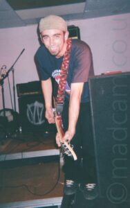 A man wearing a reversed sandy-brown cap on his head with dark sideburns underneath, as well as a dark goatee beard, also wearing a dark blue t-shirt with an indistinct white, purple, and red logo on the front, and black trousers. A red, white, and black guitar strap hangs over his left shoulder, supporting the brown electric guitar he is playing. He is hunched over slightly, and grinning at the camera. A tall black speaker stands on the floor immediately to his left, with another speaker towards the rear, with a white "W" logo in the outline of a white circle on the front grill. White walls and grey ceiling fill the rest of the frame.