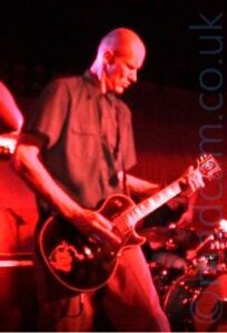 A tall man with a shaven head, wearing a dark coloured short-sleeved shirt and dark trousers, playing a black electric guitar, looking down at his left hand on the fretboard. Behind him, a man can be seen sitting behind a drum kit ion the bottom right corner. Red light floods the rest of the image.