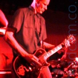 A tall man with a shaven head, wearing a dark coloured short-sleeved shirt and dark trousers, playing a black electric guitar, looking down at his left hand on the fretboard. Behind him, a man can be seen sitting behind a drum kit ion the bottom right corner. Red light floods the rest of the image.