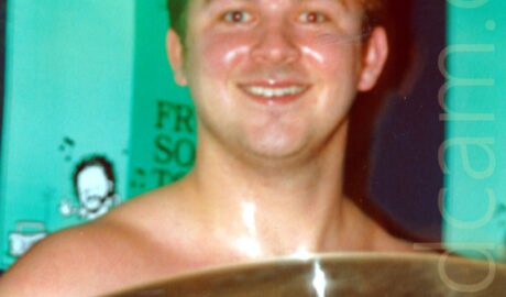A rather sweaty man with sort dark hair and his top off, sitting behind a drum kit and grinning at the camera. A large cymbal hangs in front of him, while green posters fill the wall behind him.