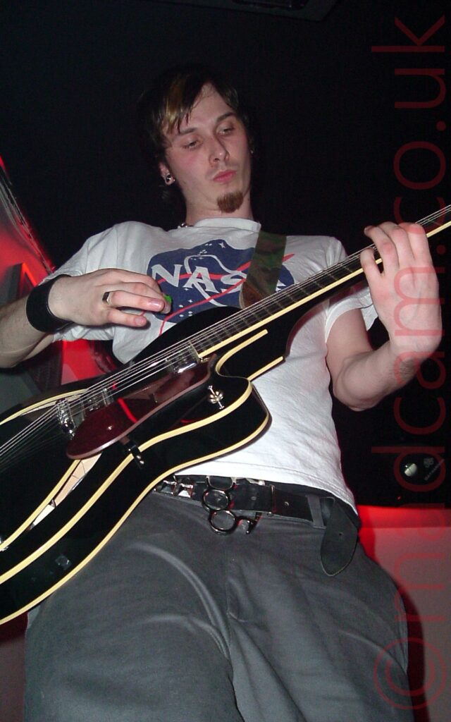 A low shot of a man with dark hair, wearing a white t-shirt with a blue, white, and red "Nasa" logo on the front, and grey trousers, playing a black electric guitar, looking down at his left hand. Red lights highlight the grey walls in the background, with black ceiling filling the rest of the frame.