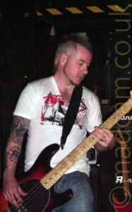 A man with short, spiky blond hair, wearing a white t-shirt with a red and black logo across the chest, partially obscured by the black strap that supports the black bass guitar he is playing. His arms are heavily tattooed. Black walls and ceiling fill the background, with black and yellow hazard tape on the wall at the top of the frame.