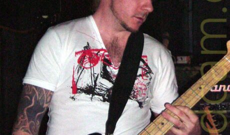 A man with short, spiky blond hair, wearing a white t-shirt with a red and black logo across the chest, partially obscured by the black strap that supports the black bass guitar he is playing. His arms are heavily tattooed. Black walls and ceiling fill the background, with black and yellow hazard tape on the wall at the top of the frame.