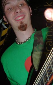 Low-down close-up of the head and shoulders of a man with lank dark hair sticking to the side of his face, a brown goatee beard, and a silvery metal ring piercing his bottom right lip. He is sweating profusely, and has a wide-eyed rictus grin on his face. He is wearing a necklace made up of metal ball on a metal chain, and a green t-shirt with a circular red logo in the middle of the chest. He is playing a black electric guitar which is attached to a camouflage-pattern guitar strap slung over his left shoulder Black ceiling fills the background, with a strip of black and yellow hazard tape running along the left side.