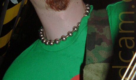 Low-down close-up of the head and shoulders of a man with lank dark hair sticking to the side of his face, a brown goatee beard, and a silvery metal ring piercing his bottom right lip. He is sweating profusely, and has a wide-eyed rictus grin on his face. He is wearing a necklace made up of metal ball on a metal chain, and a green t-shirt with a circular red logo in the middle of the chest. He is playing a black electric guitar which is attached to a camouflage-pattern guitar strap slung over his left shoulder Black ceiling fills the background, with a strip of black and yellow hazard tape running along the left side.