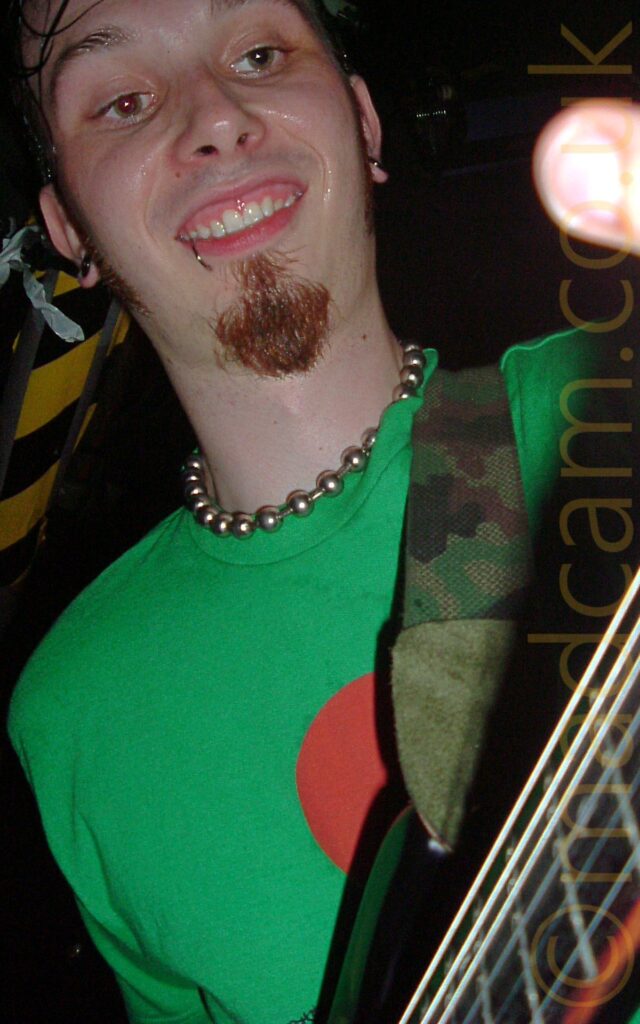 Low-down close-up of the head and shoulders of a man with lank dark hair sticking to the side of his face, a brown goatee beard, and a silvery metal ring piercing his bottom right lip. He is sweating profusely, and has a wide-eyed rictus grin on his face. He is wearing a necklace made up of metal ball on a metal chain, and a green t-shirt with a circular red logo in the middle of the chest. He is playing a black electric guitar which is attached to a camouflage-pattern guitar strap slung over his left shoulder Black ceiling fills the background, with a strip of black and yellow hazard tape running along the left side.