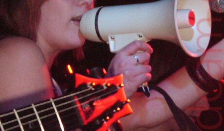 A woman, facing to the right, with her red hair cut in a bob, holding a white megaphone to her mouth, against a microphone on a stand. She is wearing a brown top with thin cream stripes, that leaves her arms and shoulders bare. The head of a black electric guitar comes in to the foreground from the left, while a man wearing a green t-shirt stands in the background on the right. Black walls and ceiling fill the top of the frame, with hints of a red light in the top right corner.