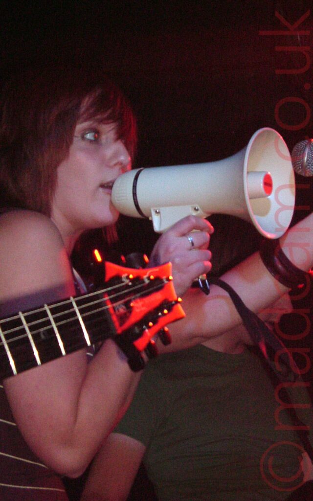 A woman, facing to the right, with her red hair cut in a bob, holding a white megaphone to her mouth, against a microphone on a stand. She is wearing a brown top with thin cream stripes, that leaves her arms and shoulders bare. The head of a black electric guitar comes in to the foreground from the left, while a man wearing a green t-shirt stands in the background on the right. Black walls and ceiling fill the top of the frame, with hints of a red light in the top right corner.