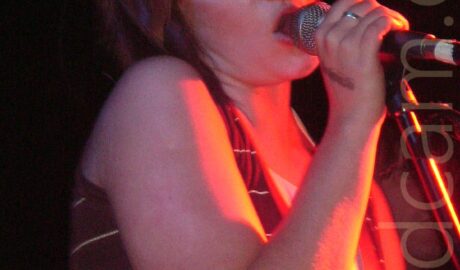 A woman with her red hair cut in a chin-length bob, wearing a brown vest top with thin yellow bands across the body, singing into a microphone on a stand she is holding in her right hand, with her head tilted slightly to her right. A red light is shining from off to the right of the frame, giving her a slight orange glow. Black walls and ceiling fill the rest of the background.