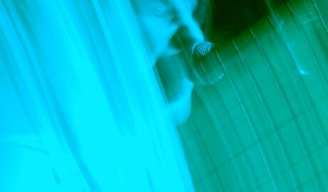 A woman performing on a stage, as part of a band. She has short dark hair, and is wearing a black and white banded top, and is singing into a microphone on a stand in front of her face. A flash has frozen her in place, while a long exposure and the stage lights have created diagonal smears of blue and green light across the frame, almost washing out everything except the black background.