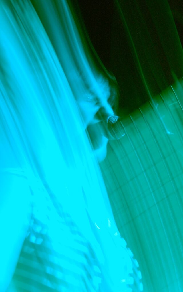 A woman performing on a stage, as part of a band. She has short dark hair, and is wearing a black and white banded top, and is singing into a microphone on a stand in front of her face. A flash has frozen her in place, while a long exposure and the stage lights have created diagonal smears of blue and green light across the frame, almost washing out everything except the black background.