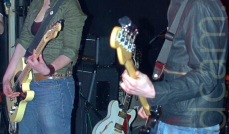 Two people on a stage as part of a band. On the left is a woman with shoulder-length red hair, wearing a green jacket over a low-cut black top and a slightly flared blue pair of jeans, playing a yellow electric guitar, and staring at a microphone on a stand in front of her, facing slightly to the left. On the right is a man with lank brown hair, wearing a black leather jacket and blue jeans,, playing a black bass guitar, while facingvto the left. A stack of speaker cabinets can be seen on the floor behind them, next to a white electric guitar. Black walls and ceiling fill the background, with red "Roadhouse" text on the wall behind them.