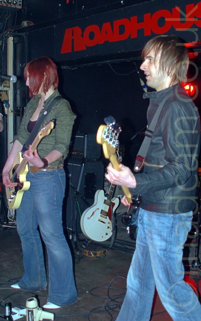 Two people on a stage as part of a band. On the left is a woman with shoulder-length red hair, wearing a green jacket over a low-cut black top and a slightly flared blue pair of jeans, playing a yellow electric guitar, and staring at a microphone on a stand in front of her, facing slightly to the left. On the right is a man with lank brown hair, wearing a black leather jacket and blue jeans,, playing a black bass guitar, while facingvto the left. A stack of speaker cabinets can be seen on the floor behind them, next to a white electric guitar. Black walls and ceiling fill the background, with red "Roadhouse" text on the wall behind them.