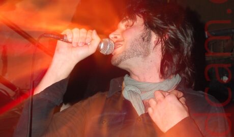 A man singing on a stage as part of a bands. He has chin-length dark hair and a close-trimmed beard, wearing a black denim shirt with a grey neckerchief around his neck, holding a microphone in his right hand, raised to his mouth as he sings, with his left hand up by his shoulder. Another person can be seen behind him on the left, wearing a grey and black banded t-shirt with a grey neckerchief tied around his neck. A flash from the camera has frozen the singer in place, while a long exposure and snap zoom in has created secondary images of the singer, that are little more than orange blooms of light on his face and body. Black walls and ceiling fill the rest of the frame.