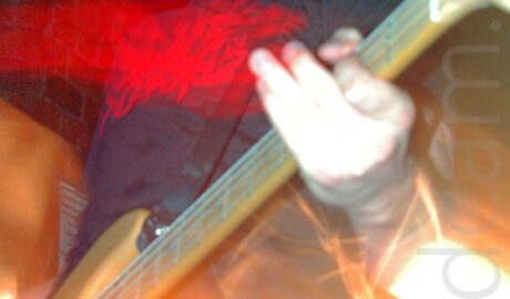 A man with short dark hair, wearing a black t-shirt with an indistinct red logo on the front, and playing a brown electric guitar, on a stage.A flash from the camera has frozen him in place, while a long exposure and his movements and the stage lights to create mostly orange secondary images of him and his instrument. Black walls and ceiling fill the rest of the frame.