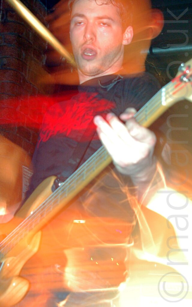 A man with short dark hair, wearing a black t-shirt with an indistinct red logo on the front, and playing a brown electric guitar, on a stage.A flash from the camera has frozen him in place, while a long exposure and his movements and the stage lights to create mostly orange secondary images of him and his instrument. Black walls and ceiling fill the rest of the frame.