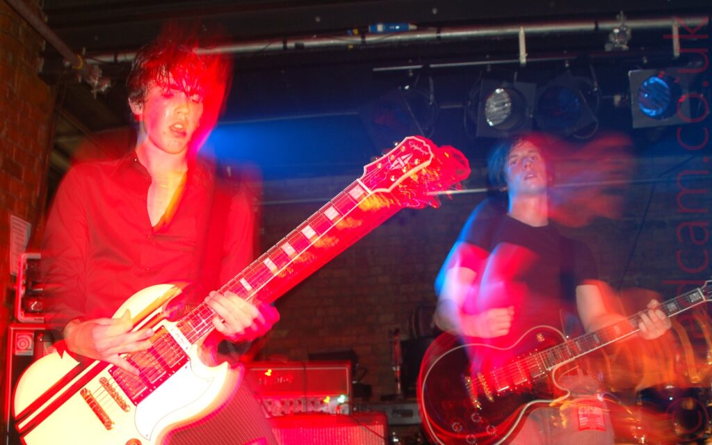 Two men playing electric guitars on a stage as part of a rock band. On the left is a man with short dark hair, wearing a black shirt with the top couple of buttons open, and black pin-stripe trousers, playing a white guitar with a pair of black stripes near the top. On the right is a man with slightly longer, dark hair, wearing a black t-shirt and faded blue jeans, playing a black electric guitar. Both men have been frozen in place by a flash from the camera, while their movements combine with a long exposure and stage lights to create secondary images of them, with booms of mostly red light around them and their instruments. Red brick walls and black ceilings fill the background, with stage lights hanging from long metal poles.