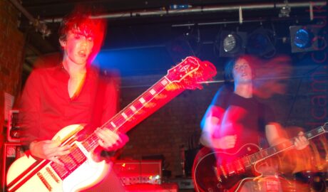 Two men playing electric guitars on a stage as part of a rock band. On the left is a man with short dark hair, wearing a black shirt with the top couple of buttons open, and black pin-stripe trousers, playing a white guitar with a pair of black stripes near the top. On the right is a man with slightly longer, dark hair, wearing a black t-shirt and faded blue jeans, playing a black electric guitar. Both men have been frozen in place by a flash from the camera, while their movements combine with a long exposure and stage lights to create secondary images of them, with booms of mostly red light around them and their instruments. Red brick walls and black ceilings fill the background, with stage lights hanging from long metal poles.