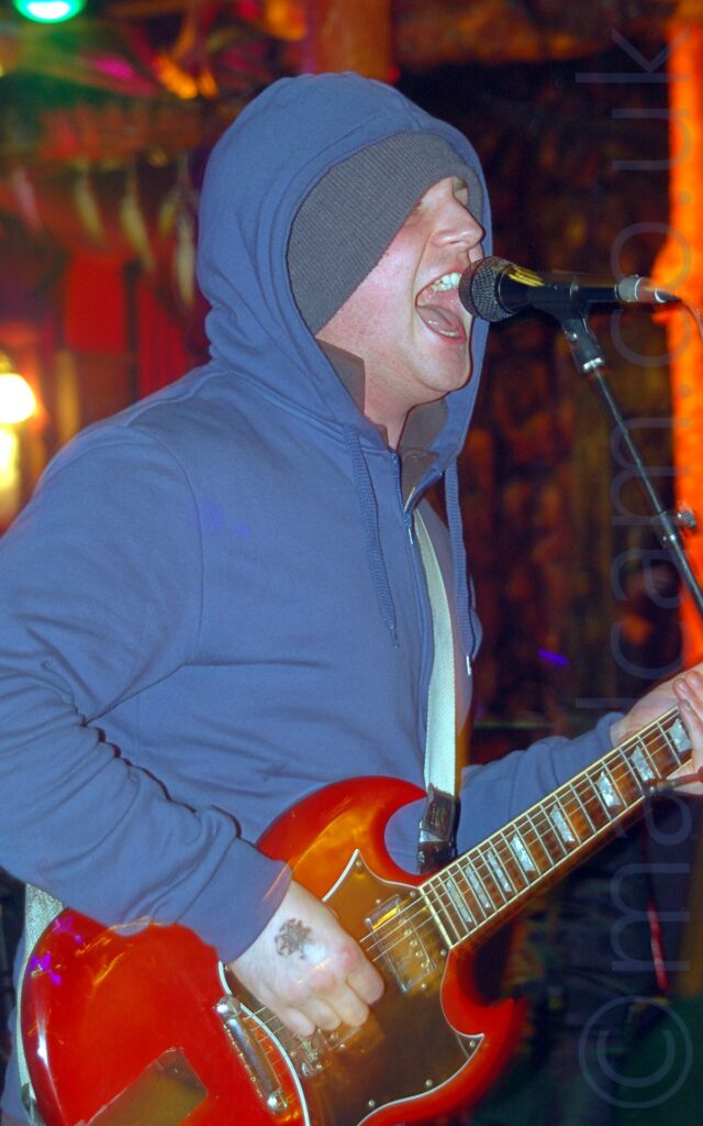 A man standing facing to the right, wearing a blue hoodie with a zip down the front, the hood pulled over his head, which also has a grey woollen hat on. He is playing a red electric guitar, attached to a pale blue strap over his left shoulder, and is singing into a microphone on a stand in front of him. An amber coloured metal pole is in the background on the right side of the frame, with another, similar pole further back, behind his head. The rest of the background is little more than blurred multi-coloured smears, with a green light peeking into frame in the top left corner.
