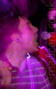A man with short dark hair, wearing a black stripey polo shirt with his head turned to his left, singing into a microphone on a stand that is leaning into the frame from the right, while holding a drumstick in his right hand, raised to his shoulder. He has a tunnel in his right earlobe. Purple stage lights and a snap zoom flood the frame with colour and streaks of light.