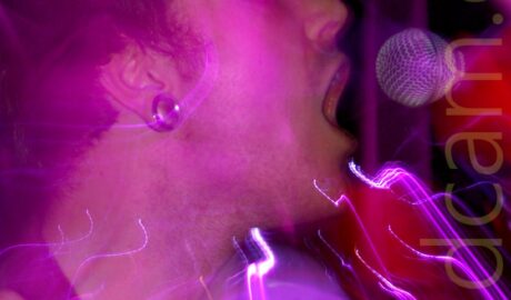A man with short dark hair, wearing a black stripey polo shirt with his head turned to his left, singing into a microphone on a stand that is leaning into the frame from the right, while holding a drumstick in his right hand, raised to his shoulder. He has a tunnel in his right earlobe. Purple stage lights and a snap zoom flood the frame with colour and streaks of light.