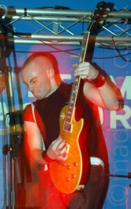 A man with short, almost spiky hair, wearing a black t-shirt, and playing an amber wood electric guitar, looking down towards the bottom left corner, his instrument partially raised in the air. A blue vinyl sign fills the background, with undecipherable script on the middle, and a metal frame holding up stage lights stretching across the top of the frame. A camera flash has frozen him in place, but his movements, combined with the stage lights, have created orange and red secondary images of him.