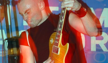 A man with short, almost spiky hair, wearing a black t-shirt, and playing an amber wood electric guitar, looking down towards the bottom left corner, his instrument partially raised in the air. A blue vinyl sign fills the background, with undecipherable script on the middle, and a metal frame holding up stage lights stretching across the top of the frame. A camera flash has frozen him in place, but his movements, combined with the stage lights, have created orange and red secondary images of him.