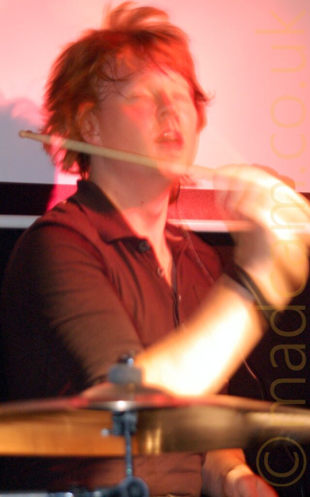A man with floppy brown hair, wearing a black shirt, sitting behind a drum kit, with his right hand holding a drumstick in the air as he faces to the right of the frame. A long exposure captures his movements, especially on his arm and hair. A brass cymbal fills the foreground, with white walls coloured salmon pink by stage lights filling the background.