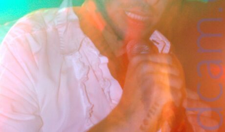 Close-up of a man with shaggy dark hair, wearing a frilly white shirt, singing into a microphone held in his right hand. The words "Big shoes to fill" have been tattooed on the inside edge of his right forearm. A long exposure has made him slightly indistinct, his movement and blooms of red and green lights creating artifacts and secondary images all over the frame.
