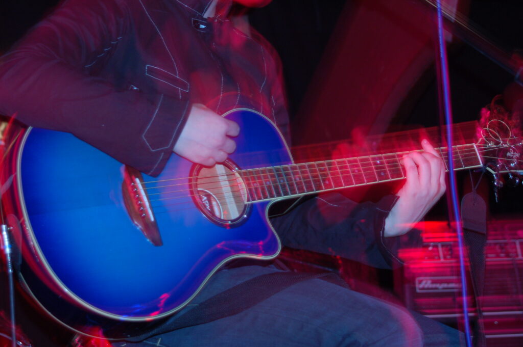 A close-up of the blue acoustic guitar being played by a member of a band performing on a stage. They are wearing a grey jacket with thin white piping on the seams, and a faded blue pair of jeans, and are sitting on a stool. A flash from the camera has frozen them in place, while a long exposure and their movements combine with the red stage lights to create secondary images and smear of light following their movements. A stack of grey and black amplifiers is in the background in the bottom right corner, with black walls and ceiling filling the rest of the frame.