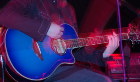 A close-up of the blue acoustic guitar being played by a member of a band performing on a stage. They are wearing a grey jacket with thin white piping on the seams, and a faded blue pair of jeans, and are sitting on a stool. A flash from the camera has frozen them in place, while a long exposure and their movements combine with the red stage lights to create secondary images and smear of light following their movements. A stack of grey and black amplifiers is in the background in the bottom right corner, with black walls and ceiling filling the rest of the frame.